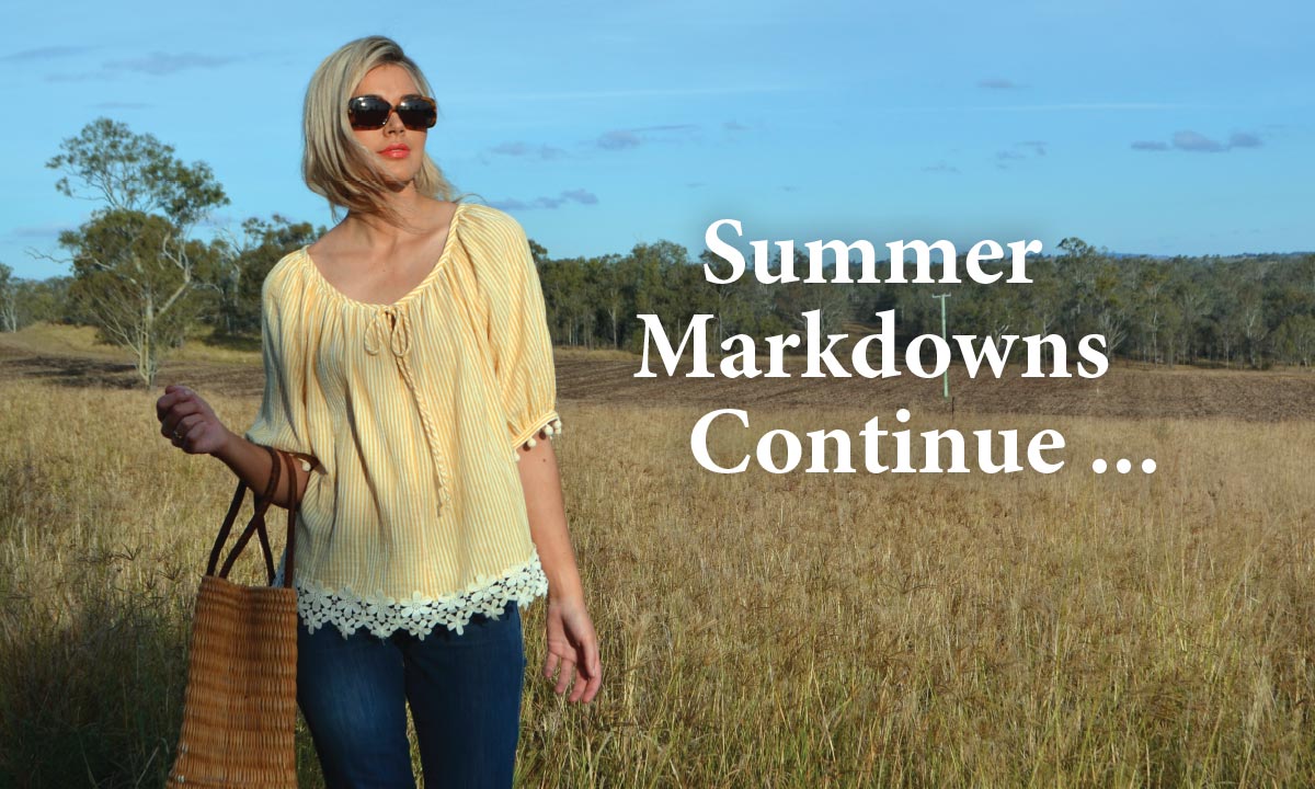 Model wearing Daisy Chain Blouse standing in a grassy field and Australian landscape in the Lockyer Valley in Queensland. She is wearing sunglasses. She is to the left of the image, with negative space to the right, with text that reads 'Summer Markdowns Continue' reversed out over the landscape. Click this image to view the products available.